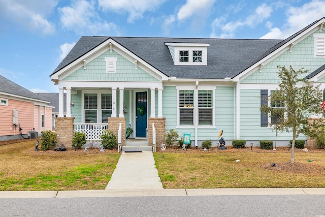 craftsman-style home featuring cooling unit, covered porch, a front lawn, and a shingled roof