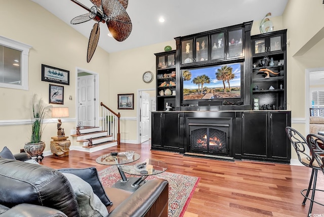 living area with vaulted ceiling, ceiling fan, wood finished floors, a warm lit fireplace, and stairs