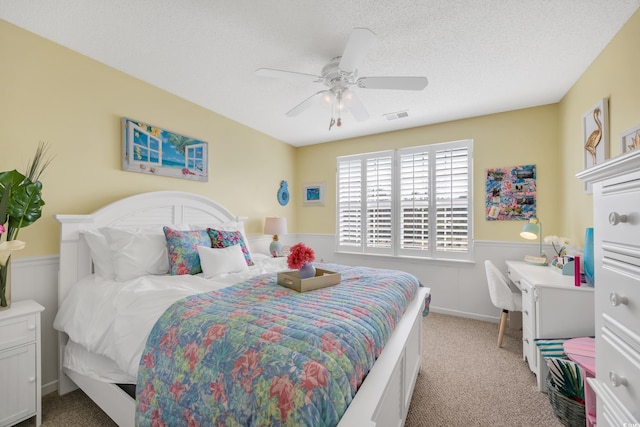 bedroom featuring light carpet, visible vents, a ceiling fan, wainscoting, and a textured ceiling