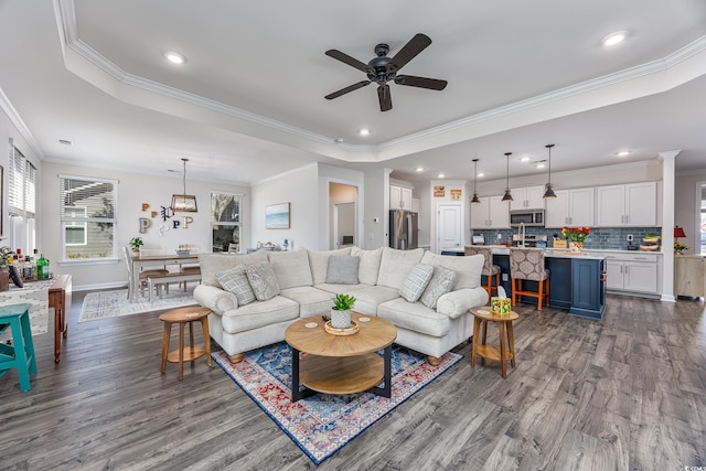 living area featuring a tray ceiling, dark wood-type flooring, ornamental molding, a ceiling fan, and baseboards