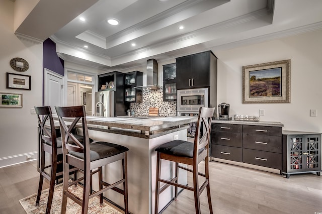 kitchen featuring a tray ceiling, a breakfast bar, tasteful backsplash, appliances with stainless steel finishes, and wall chimney range hood