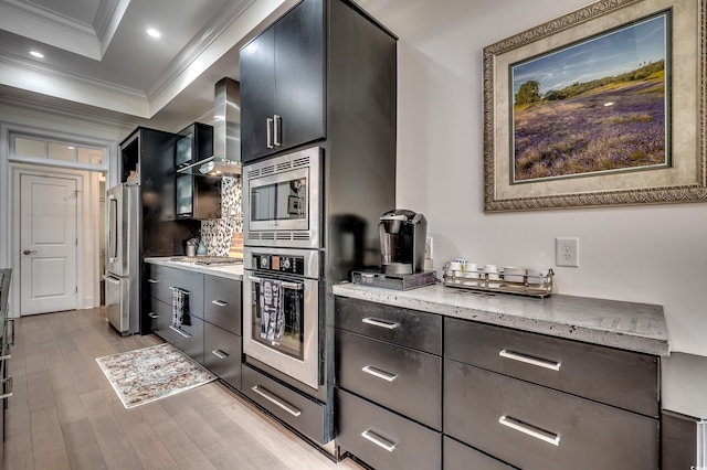 kitchen featuring a raised ceiling, appliances with stainless steel finishes, ornamental molding, wall chimney range hood, and light wood-type flooring