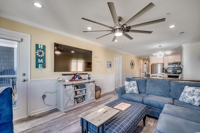 living room with crown molding, visible vents, wainscoting, light wood-type flooring, and ceiling fan with notable chandelier