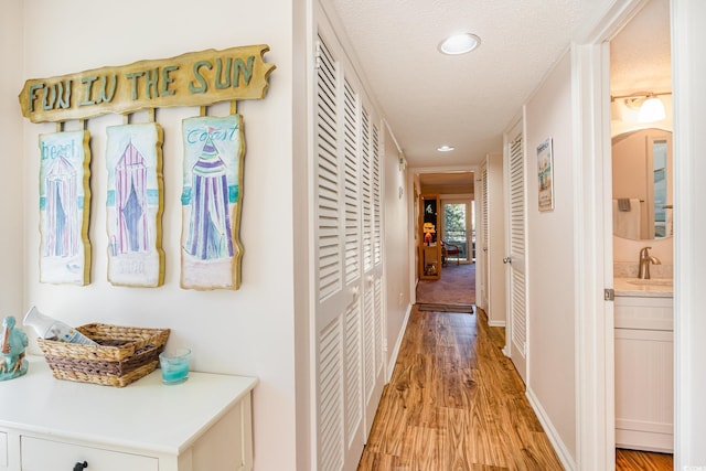 corridor featuring baseboards, light wood-style flooring, a textured ceiling, a sink, and recessed lighting