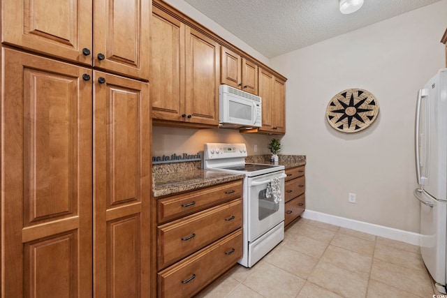 kitchen featuring brown cabinetry, a textured ceiling, dark stone counters, white appliances, and baseboards