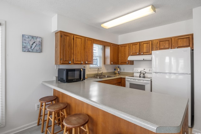 kitchen with under cabinet range hood, brown cabinets, a peninsula, white appliances, and a sink