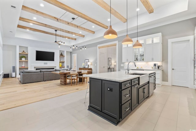 kitchen featuring white cabinets, an island with sink, glass insert cabinets, a sink, and beam ceiling