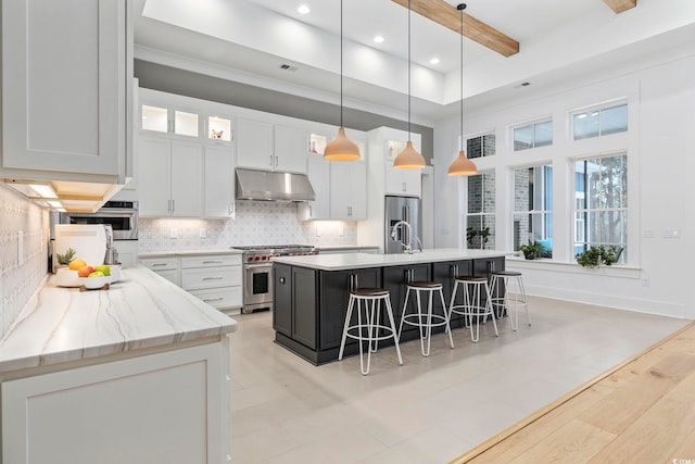 kitchen with white cabinets, appliances with stainless steel finishes, a breakfast bar, under cabinet range hood, and beam ceiling