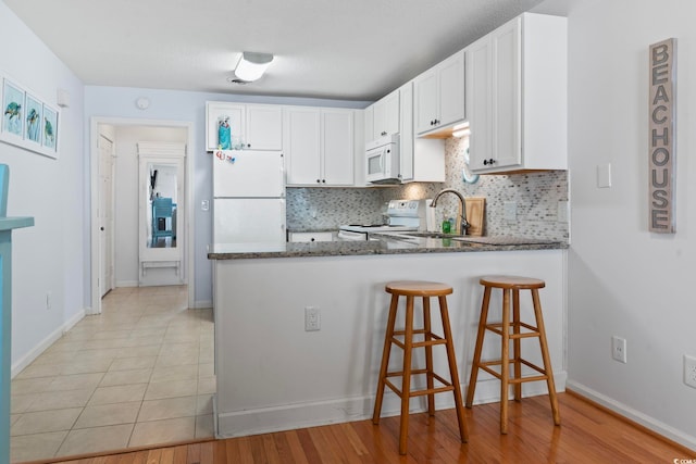 kitchen with a peninsula, white appliances, white cabinetry, backsplash, and dark stone counters