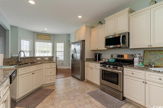 kitchen featuring appliances with stainless steel finishes, cream cabinetry, a sink, recessed lighting, and stone counters
