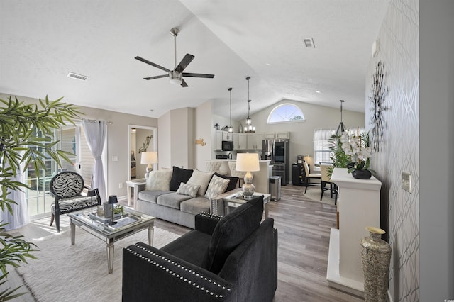 living area featuring visible vents, high vaulted ceiling, light wood-style flooring, and ceiling fan with notable chandelier