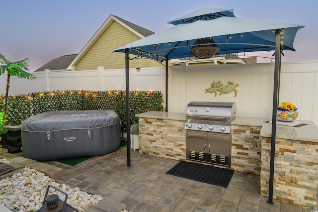 patio terrace at dusk featuring grilling area, a hot tub, an outdoor kitchen, and fence