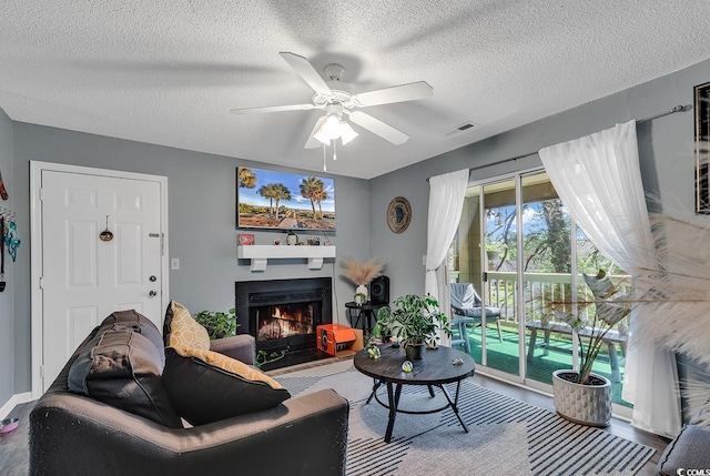 living area featuring a warm lit fireplace, visible vents, ceiling fan, and a textured ceiling