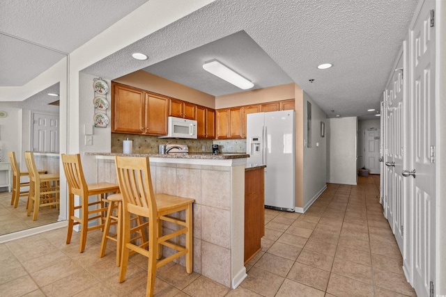 kitchen featuring decorative backsplash, brown cabinetry, white appliances, a peninsula, and a kitchen bar