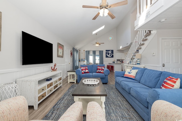 living room featuring a skylight, a ceiling fan, a wainscoted wall, wood finished floors, and stairs