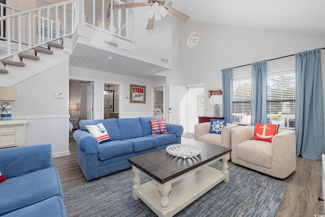 living area with a wainscoted wall, ceiling fan, wood finished floors, high vaulted ceiling, and stairs