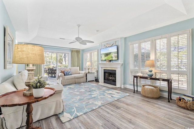 living area featuring baseboards, a fireplace with flush hearth, ceiling fan, wood finished floors, and a tray ceiling