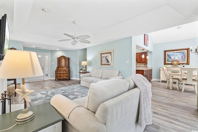 living room featuring ceiling fan with notable chandelier, a tray ceiling, light wood-style flooring, and baseboards