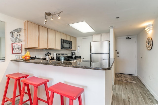 kitchen with a peninsula, appliances with stainless steel finishes, light brown cabinetry, tasteful backsplash, and dark stone countertops