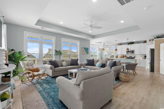 living room with a ceiling fan, visible vents, light wood finished floors, a tray ceiling, and recessed lighting