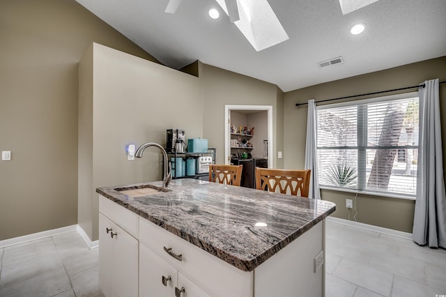 kitchen with visible vents, lofted ceiling with skylight, a sink, an island with sink, and dark stone counters