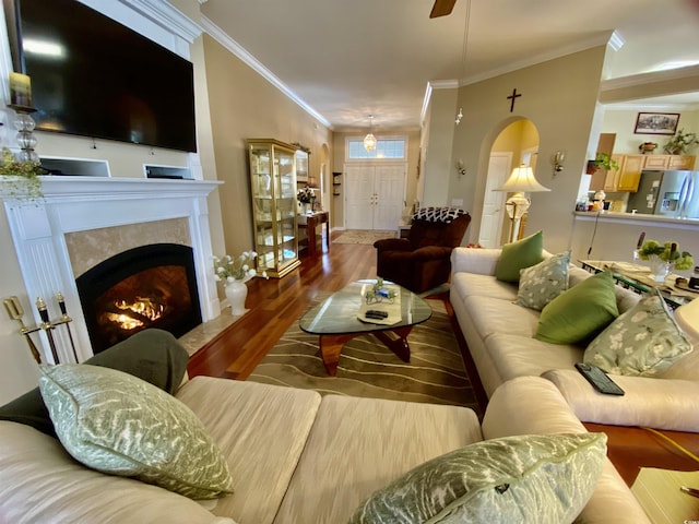 living room with arched walkways, ceiling fan, wood finished floors, a lit fireplace, and crown molding