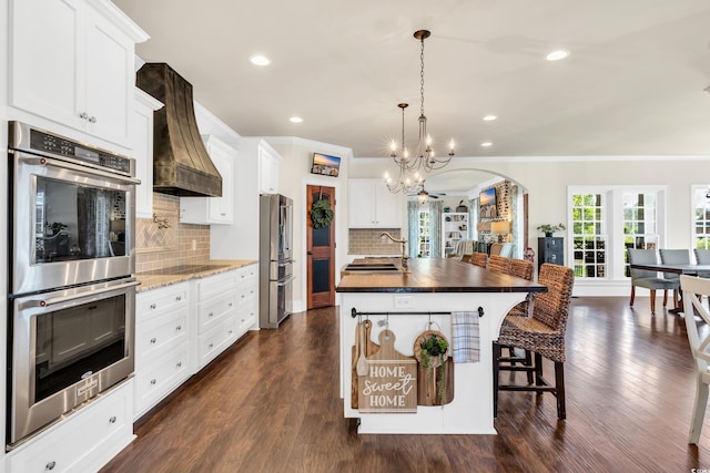 kitchen with arched walkways, stainless steel appliances, premium range hood, ornamental molding, and dark wood finished floors