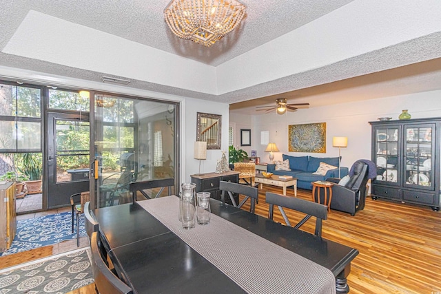 dining room with a textured ceiling, light wood finished floors, ceiling fan with notable chandelier, and a raised ceiling