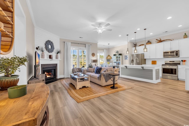 living area with ceiling fan, light wood finished floors, a fireplace with flush hearth, and crown molding