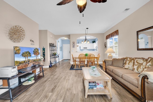 living area featuring arched walkways, light wood-style flooring, ceiling fan with notable chandelier, visible vents, and baseboards