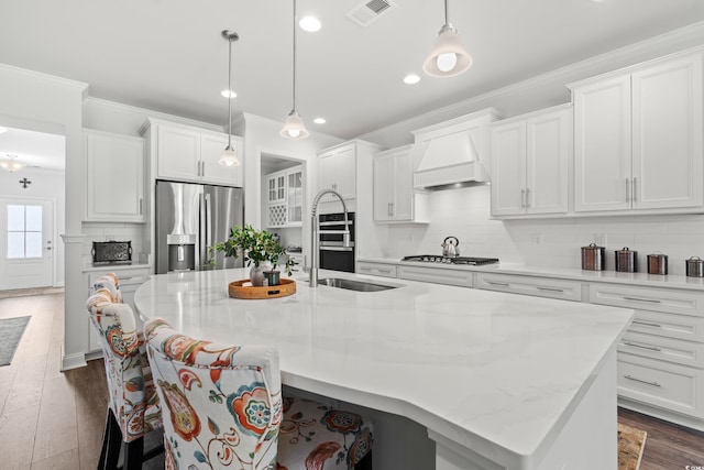 kitchen featuring an island with sink, custom range hood, ornamental molding, appliances with stainless steel finishes, and dark wood-style floors