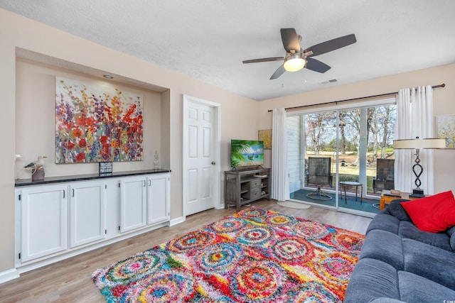 living room with light wood-type flooring, visible vents, a textured ceiling, baseboards, and ceiling fan