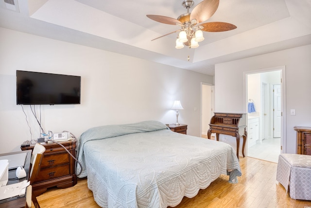 bedroom featuring visible vents, a tray ceiling, ensuite bathroom, light wood-style floors, and a ceiling fan