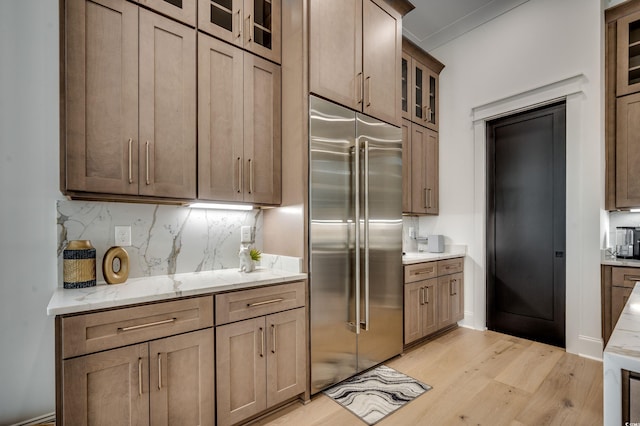 kitchen featuring light stone counters, light wood-style floors, backsplash, stainless steel built in fridge, and glass insert cabinets