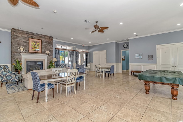 dining room featuring ceiling fan, light tile patterned flooring, a large fireplace, wainscoting, and crown molding