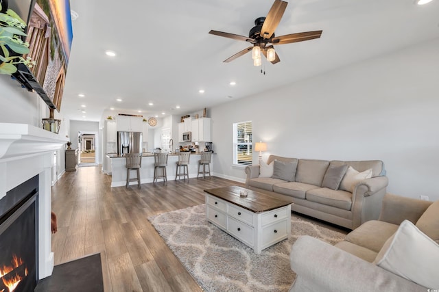 living area featuring a warm lit fireplace, baseboards, ceiling fan, light wood-type flooring, and recessed lighting