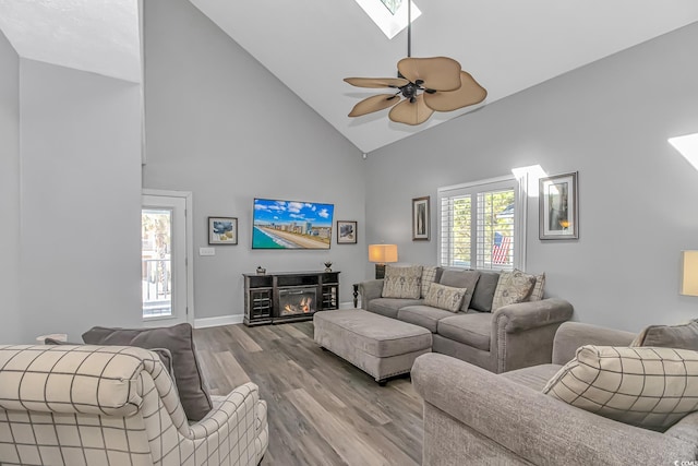 living room with a skylight, a glass covered fireplace, ceiling fan, wood finished floors, and high vaulted ceiling