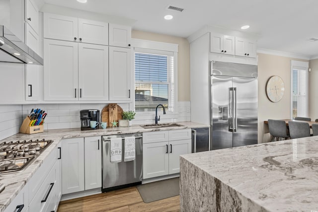 kitchen with appliances with stainless steel finishes, a healthy amount of sunlight, white cabinets, a sink, and wall chimney range hood