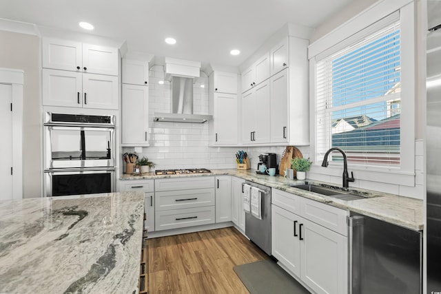 kitchen featuring light wood finished floors, wall chimney exhaust hood, a sink, stainless steel appliances, and backsplash