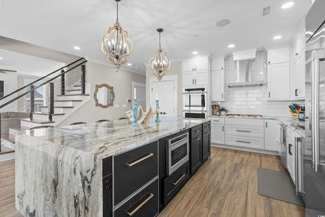 kitchen with appliances with stainless steel finishes, white cabinets, wall chimney range hood, wood finished floors, and dark cabinetry