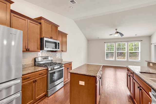 kitchen featuring light stone counters, vaulted ceiling, appliances with stainless steel finishes, backsplash, and brown cabinetry