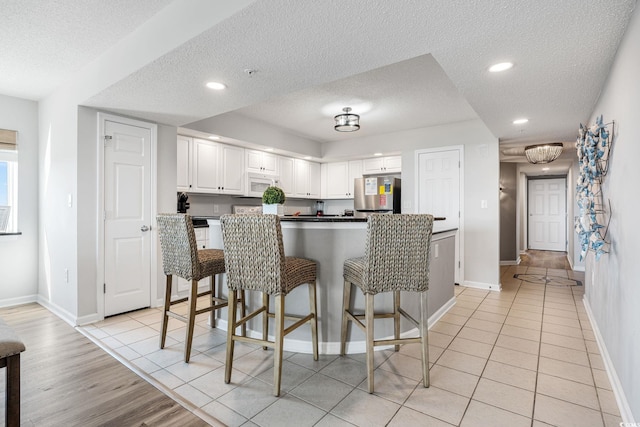 kitchen with light tile patterned floors, white microwave, freestanding refrigerator, white cabinets, and a kitchen bar