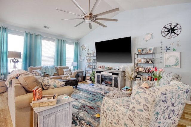 living room with a ceiling fan, visible vents, vaulted ceiling, a glass covered fireplace, and light wood-type flooring