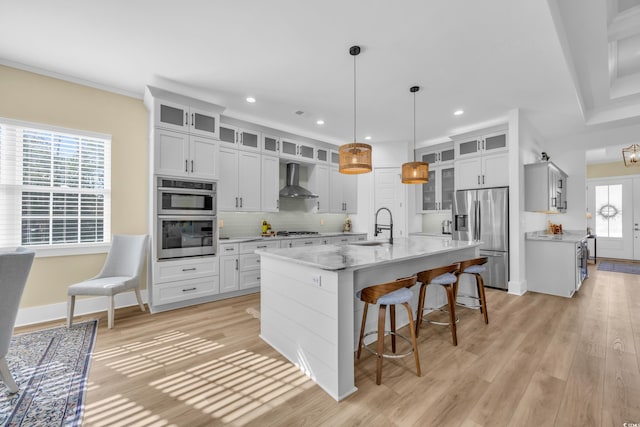 kitchen featuring a breakfast bar area, a kitchen island with sink, a sink, appliances with stainless steel finishes, and wall chimney exhaust hood