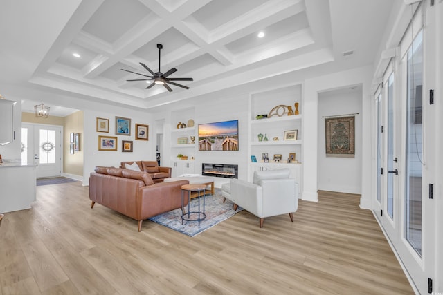 living room featuring built in shelves, coffered ceiling, ceiling fan, light wood-style floors, and a glass covered fireplace