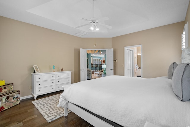 bedroom with a ceiling fan, a tray ceiling, connected bathroom, baseboards, and dark wood-style flooring