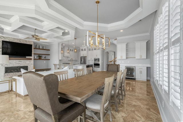 dining space with beverage cooler, coffered ceiling, ornamental molding, a stone fireplace, and ceiling fan with notable chandelier