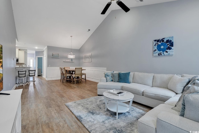 living area featuring high vaulted ceiling, light wood-style flooring, wainscoting, a decorative wall, and ceiling fan