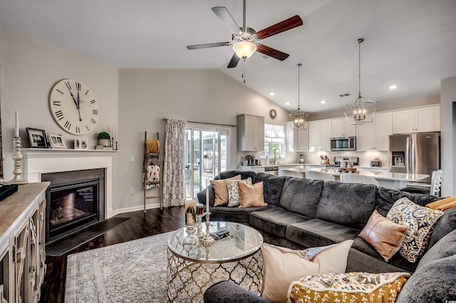 living room with vaulted ceiling, ceiling fan with notable chandelier, recessed lighting, a glass covered fireplace, and dark wood-style flooring