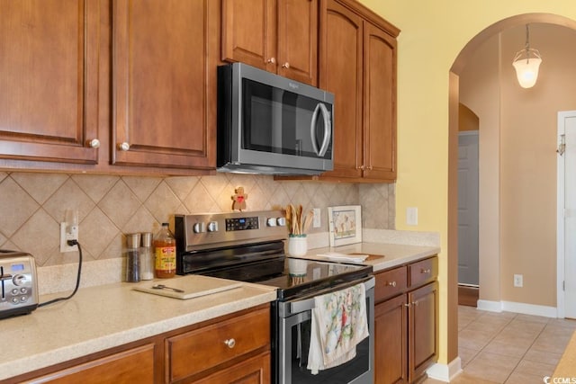 kitchen with light tile patterned floors, arched walkways, stainless steel appliances, decorative backsplash, and brown cabinets